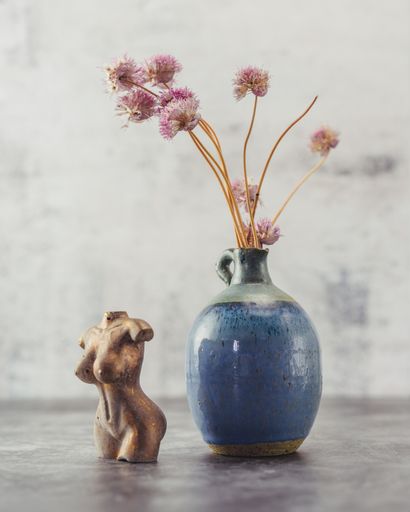 A vertical closeup of a blue vase with pinkish flowers with a woman's body figure on the gray surface.