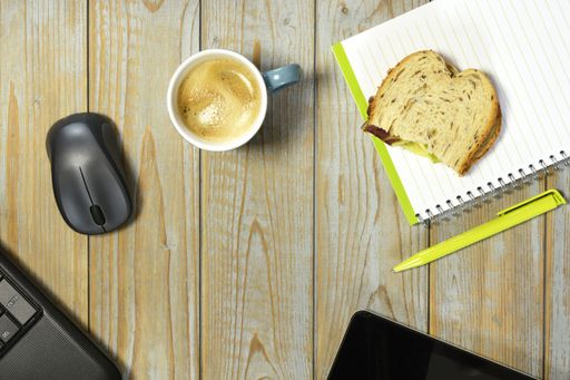 A wireless mouse, a cup of coffee, piece of bread and notebook with pen isolated on a wooden surface - work at home concept