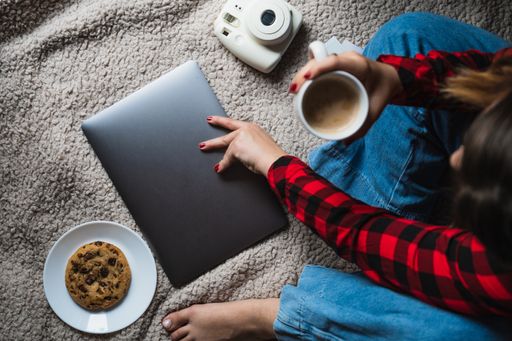 A horizontal shot of a female sitting on a blanket with her laptop, coffee and cookie