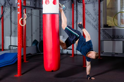 Young MMA fighter in inverted state delivers strong straight kick on red boxing pear to area of alleged head of opponent. Training process in gym.