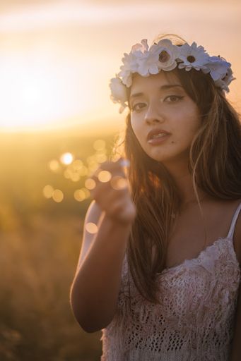 A vertical shot of a young caucasian female in a white dress and white flower wreath posing with a Bangle light in a field