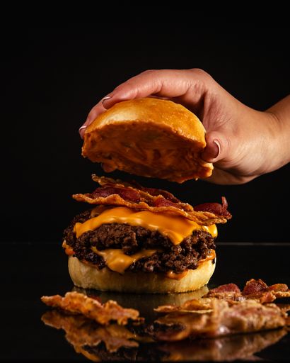 A person preparing a delicious burger with beef, yellow cheese, and bacon