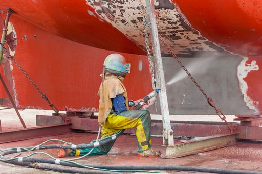 worker, sandblasting the corroded hull of a sailing vessel with a high pressure sandblasting syst