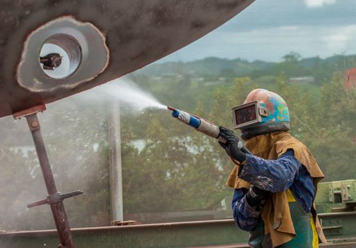 worker, sandblasting the corroded hull of a sailing vessel with a high pressure sandblasting syst