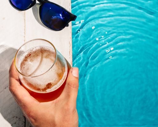 A top view of person's hand on the pool holding a glass of beer next to sunglasses