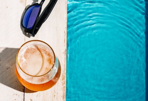 A top view of a glass of beer next to sunglasses by the pool on the coping