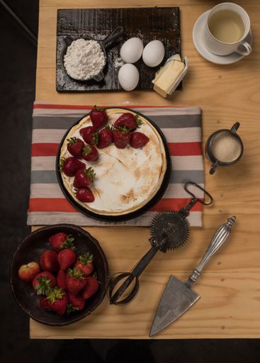 A top view of strawberry cake on wooden table with eggs, butter, flour and cup of tea