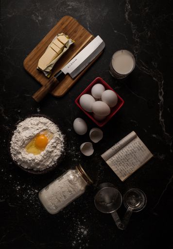 A top view of the butter, flour, eggs and a cup of milk on a black marble table