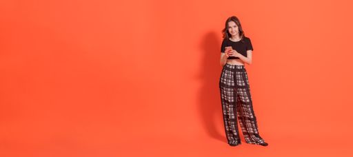 Young brunette girl in black pantsuit stands on orange background. He holds mobile phone in his hands and looks into camera. Negative space on left.