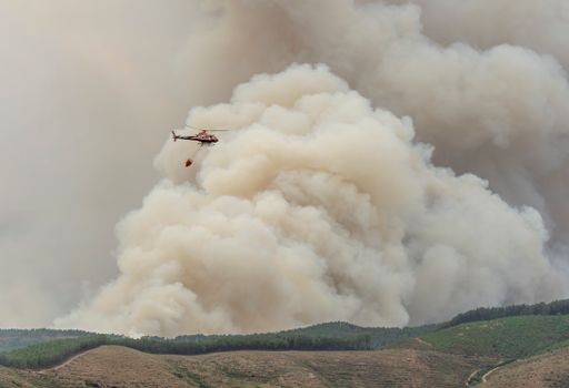 A view of a bucket dipping water to fight a fire, hanging from a helicopter against smoke
