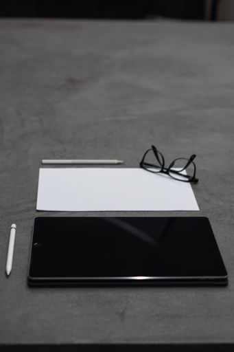A vertical closeup of a black tablet, a piece of white paper, white pencil and a pen, and eyeglasses