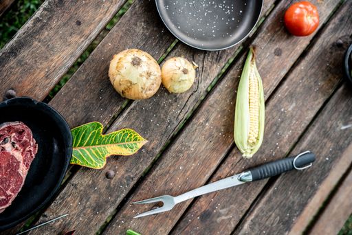 A top view of a wooden picnic table with a raw steak, onions, corn, and a barbecue fork