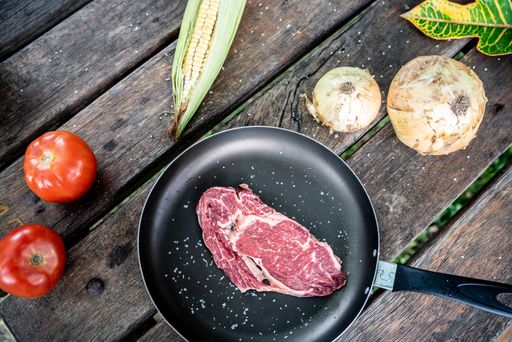 A top view of a black pan with seasoned raw steak on a wooden table with corn, tomatoes, and onions