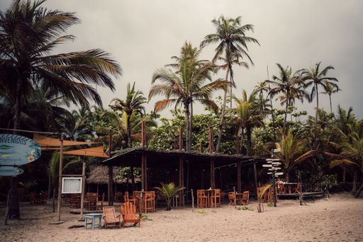 A scenic view of an empty cafe on a sandy beach against palm trees on a cloudy day