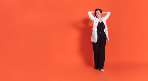 Young brunette woman in black trouser suit and white jacket stands on orange background. Raised both hands and launched them into their hair.