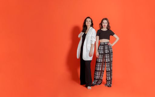 Two young women, mom and daughter, in business trouser costumes stand on orange background, smile and looking into camera.