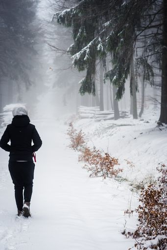 A vertical shot of a female walking on a snow covered road surrounded by trees in a winter day
