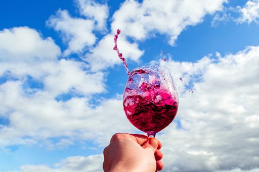 A closeup shot of a man holding a wineglass and the splashes of red wine against the sky
