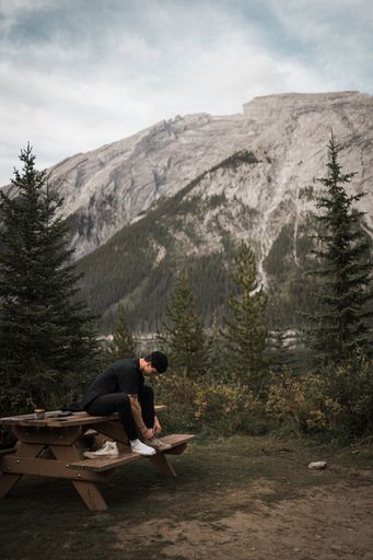 A vertical shot of a Caucasian male sitting on a bench with beautiful mountain and trees in the background