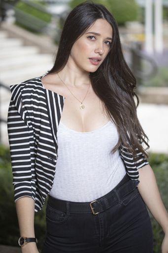 A vertical shot of an attractive female with long brown hair wearing a low neck white tanktop a striped blazer and black jeans with a belt posing