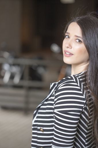 A vertical shot of an attractive brunette female wearing striped blazer leaning on a wall looking to the camera