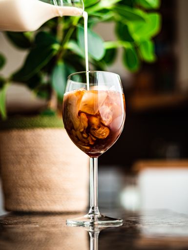 Milk being poured in a glass of iced coffee by the window on a summer day.