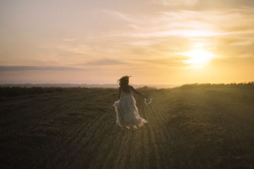 A young brunette female wearing a white dress and holding a flower crown running towards the sunset in a field