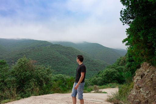 A side view of young caucasian male wearing a black shirt and jean shorts posing in front of scenic green hills on a foggy day