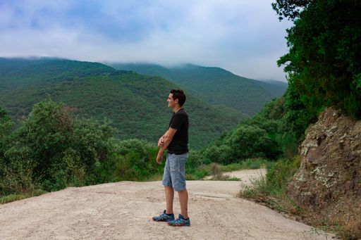 A side view of young caucasian male wearing a black shirt and jean shorts posing in front of scenic green hills on a foggy day