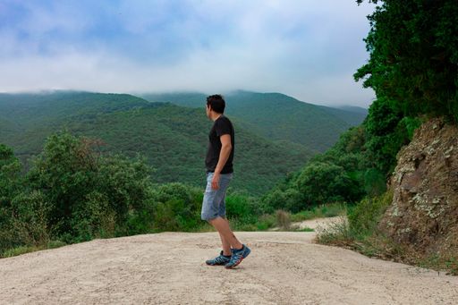 A side view of young caucasian male wearing a black shirt and jean shorts posing in front of scenic green hills on a foggy day