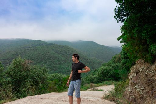 A side view of young caucasian male wearing a black shirt and jean shorts posing in front of scenic green hills on a foggy day