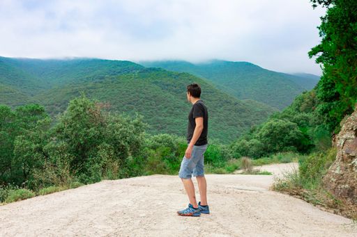 A side view of young caucasian male wearing a black shirt and jean shorts posing in front of scenic green hills on a foggy day