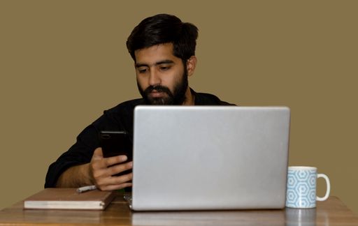 A South Asian man looking at his phone while working on a laptop