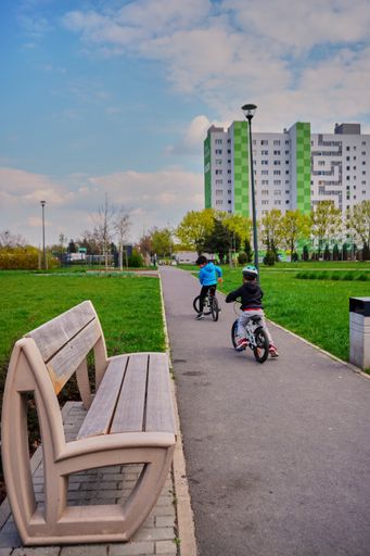 Two boys riding bikes on a footpath with park bench leading to apartment block in the Orla Bialego area, Poznan, Poland