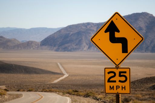 A long road in a desert with a curve sign in the foreground and some mountains in the background