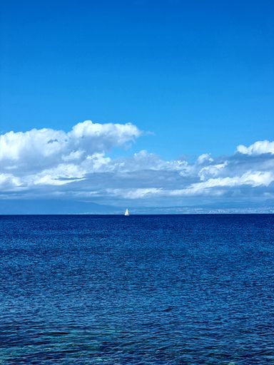 A vertical shot of the blue water of a sea with a boat sailing far away, under a cloudy, clear sky