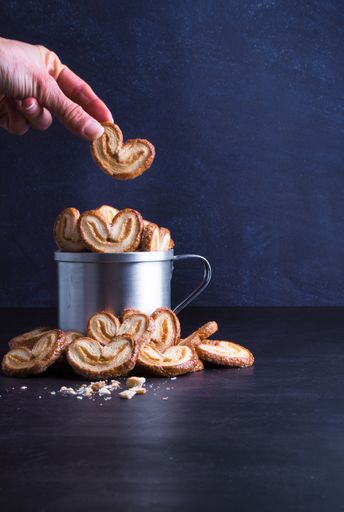 A vertical shot of a hand taking out a heart-shaped palmier pastry in from a silver jug