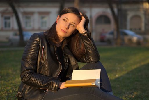 A young caucasian female sitting on the grass with a book leaning her head in her hand looking to the side and thinking
