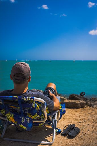 A back shot of an adult man sitting on a metal beach chair looking at the sea and blue sky in bright sunlight