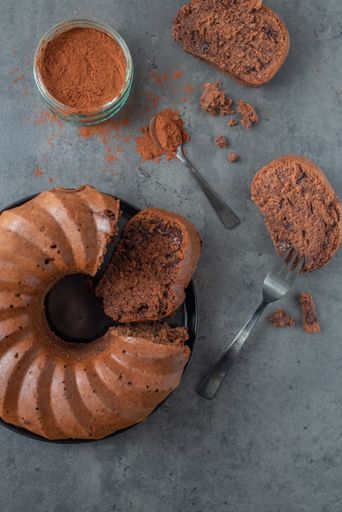 A top view of a chocolate covered pumpkin bundt cake, slices with fork and cacao powder in a jar on a gray background