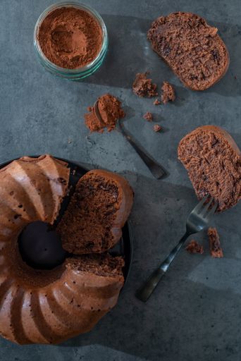 A top view of a chocolate covered pumpkin bundt cake, slices with fork and cacao powder in a jar on a gray background