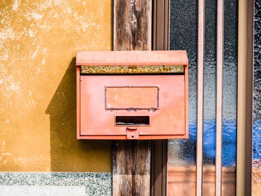 A front view of an old red mailbox attached to a yellow wall next to a window