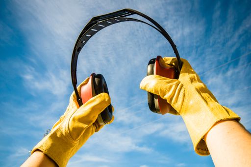 A closeup shot of yellow gloved hands holding red noise canceling construction headphones up to a blue sky background
