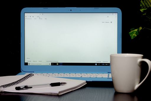 A front view of an open blue laptop, a white coffee mug and a blank notebook with a black pen
