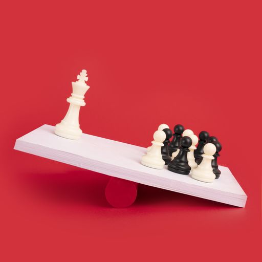 A white teeter-totter with a group of black and white chess pawns heavier than one white king piece on a red background- perfect for teamwork concepts