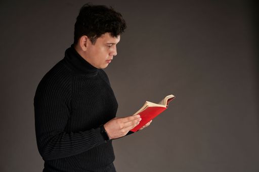 An intelligent Latin man in a black sweater reading a red book isolated on gray background