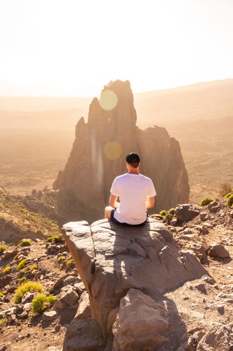 A back shot of a young tourist with a white t-shirt sitting on big rock looking at the sunset in the natural area of Teide in Tenerife, Canary Islands