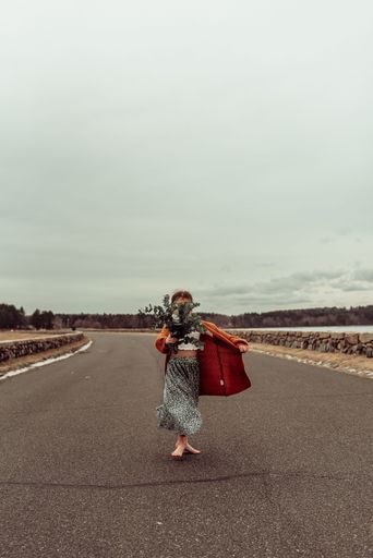 A vertical shot of a barefoot little girl holding a big bag and a bouquet of flowers on an open road