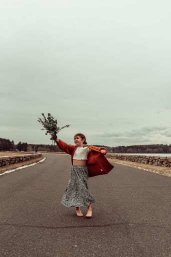 A vertical shot of a barefoot little girl holding a big bag and a bouquet of flowers on an open road