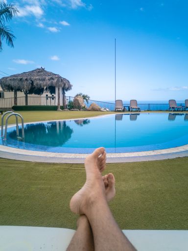 A vertical of a person's feet lying against a swimming pool reflecting beach chairs and an alcove